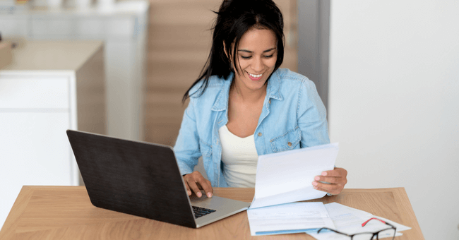 A woman works on her laptop while looking at a piece of paper.